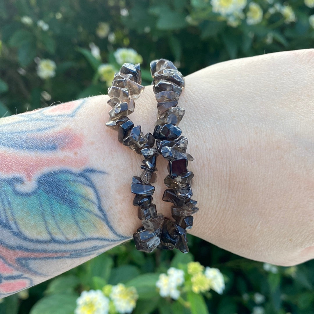 A bracelet made of smokey quartz chips displayed on a person's wrist with a blurred background of greenery.